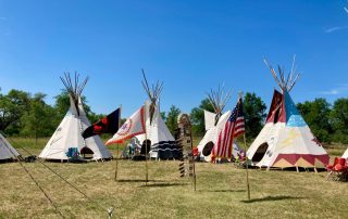 Eagle feather staff, Rosebud Sioux Tribe Flag, POW flag and U.S. flag in front of nine teepees.