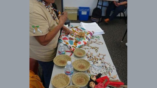 In this picture while answering questions over historical aspects of Miwok life one can see the many artistic displays that Tribal Elder Mildred Burley created and brought with her to the presentation. Traditional dolls with pine needle bodies and oak ball heads.nVarious jewelry items utilizing shells, beads, bone, acorns and feathers. Traditional miwok "Pulak-a" miwok word for basket and open scarf showing the hand game sticks used earlier while playing "hand game". Also on far edge of table were brief history handouts that were distributed to all those whom attended.